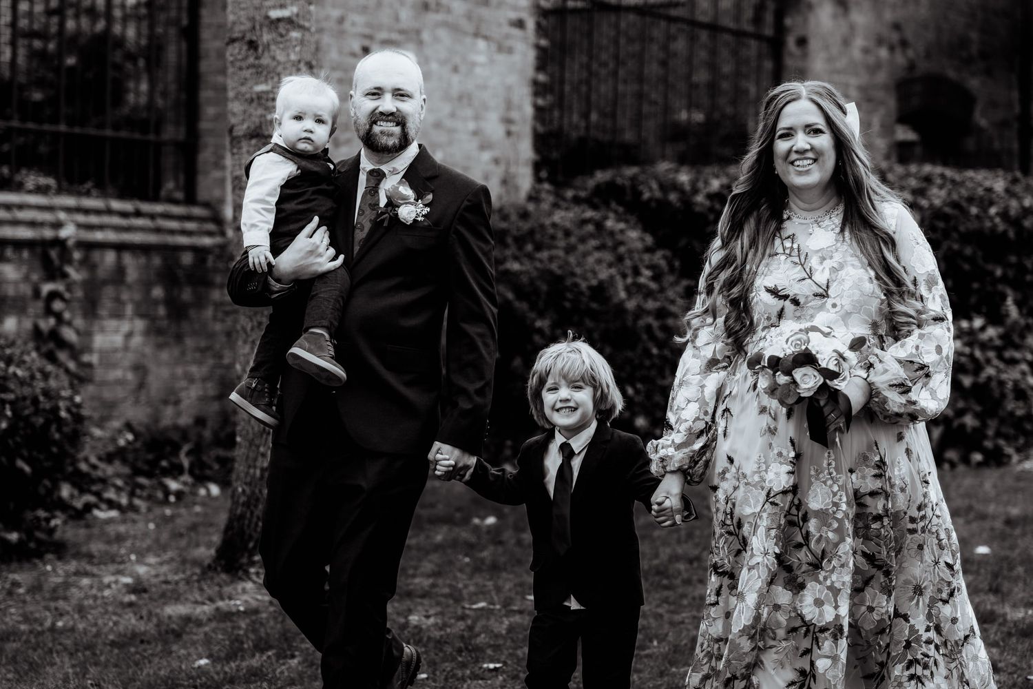 Black and white wedding photo of a couple with two young children walking outdoors near a stone building.