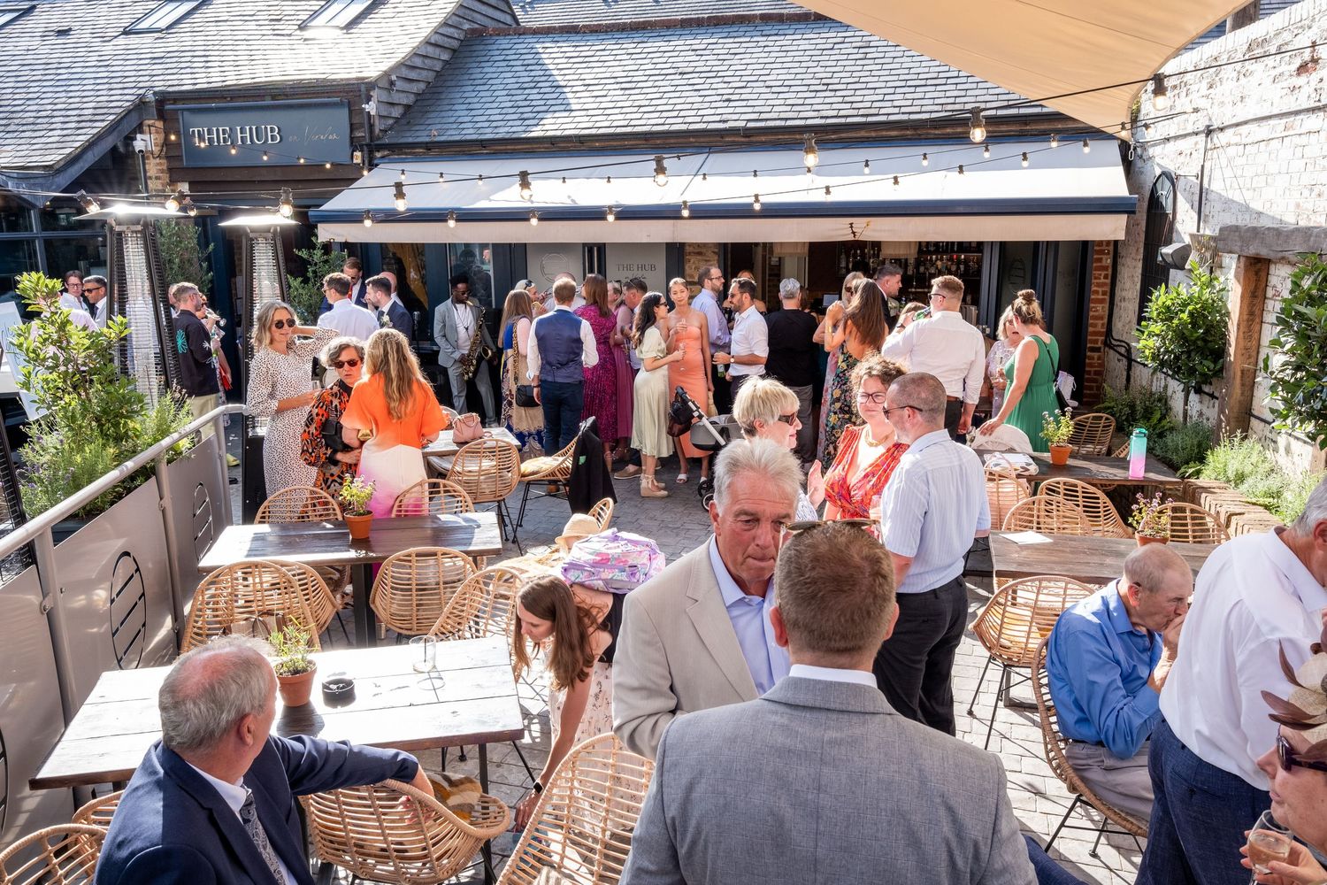 Busy outdoor patio of a restaurant with guests socializing on a sunny day, featuring rattan chairs and tables.