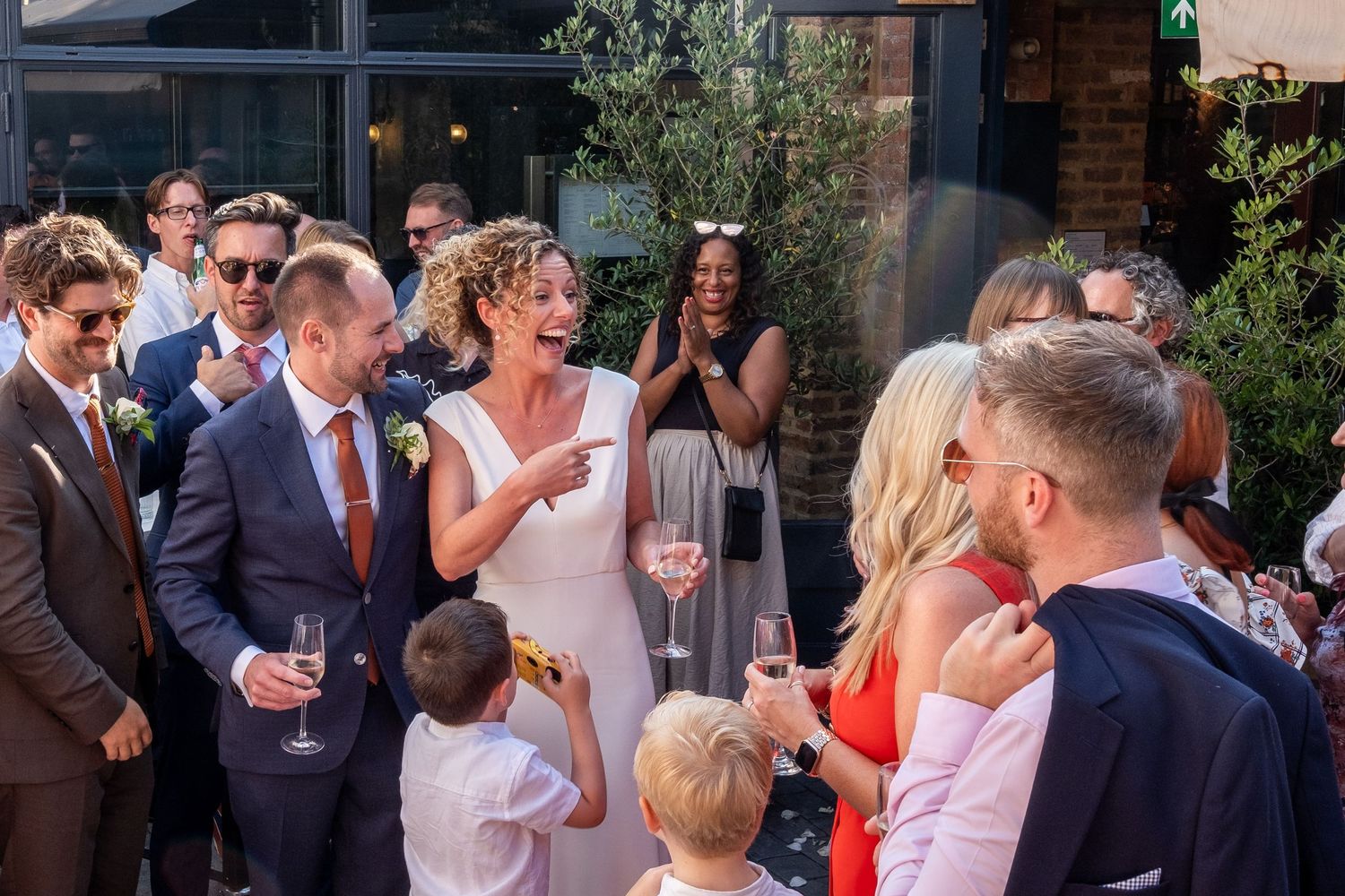 Bride in white dress laughing with guests at outdoor wedding reception, surrounded by smiling attendees holding champagne glasses.