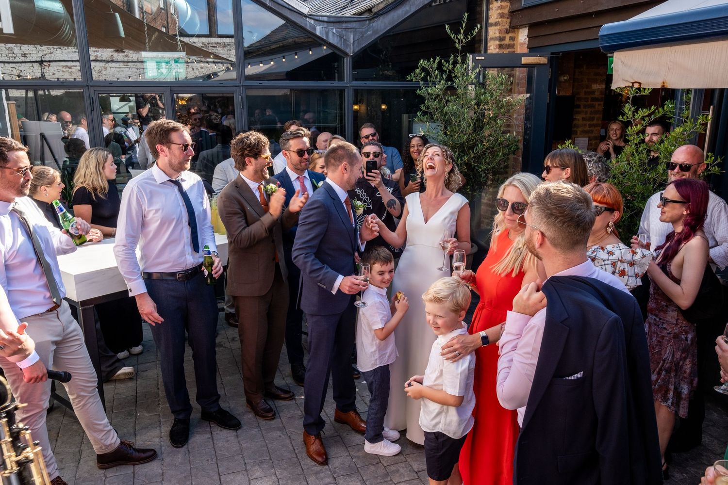 Wedding guests mingle outdoors at a sunny reception venue with the bride in a white dress.