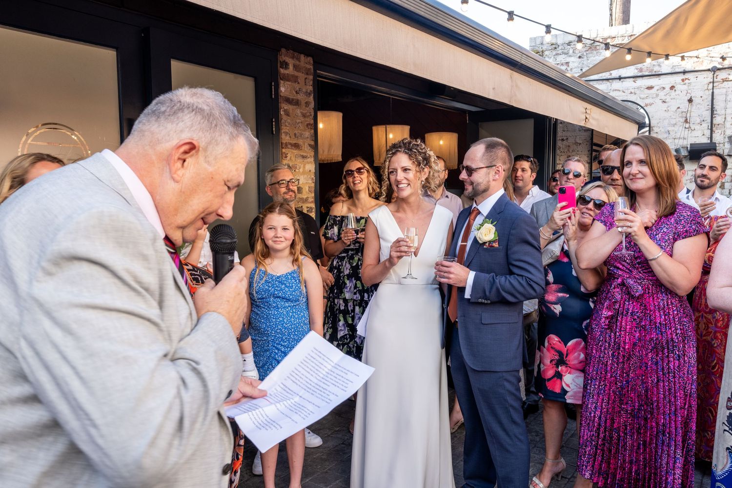 Officiant reads vows as bride and groom stand together during outdoor wedding ceremony with guests applauding.