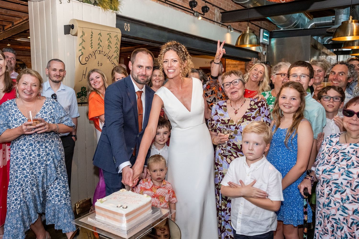 Bride and groom cutting wedding cake surrounded by smiling guests at a warm indoor wedding reception celebration.