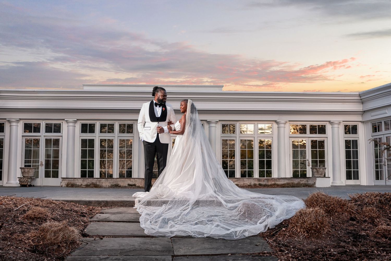 Bride and groom stand together at sunset outside an elegant white venue, her lace train flowing across steps. Deerfield Golf Club Newark De