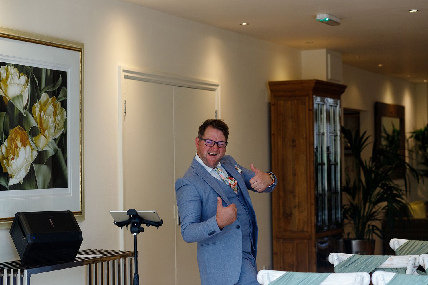 Man in light blue suit giving thumbs up at elegant indoor venue with floral artwork and green plants.
