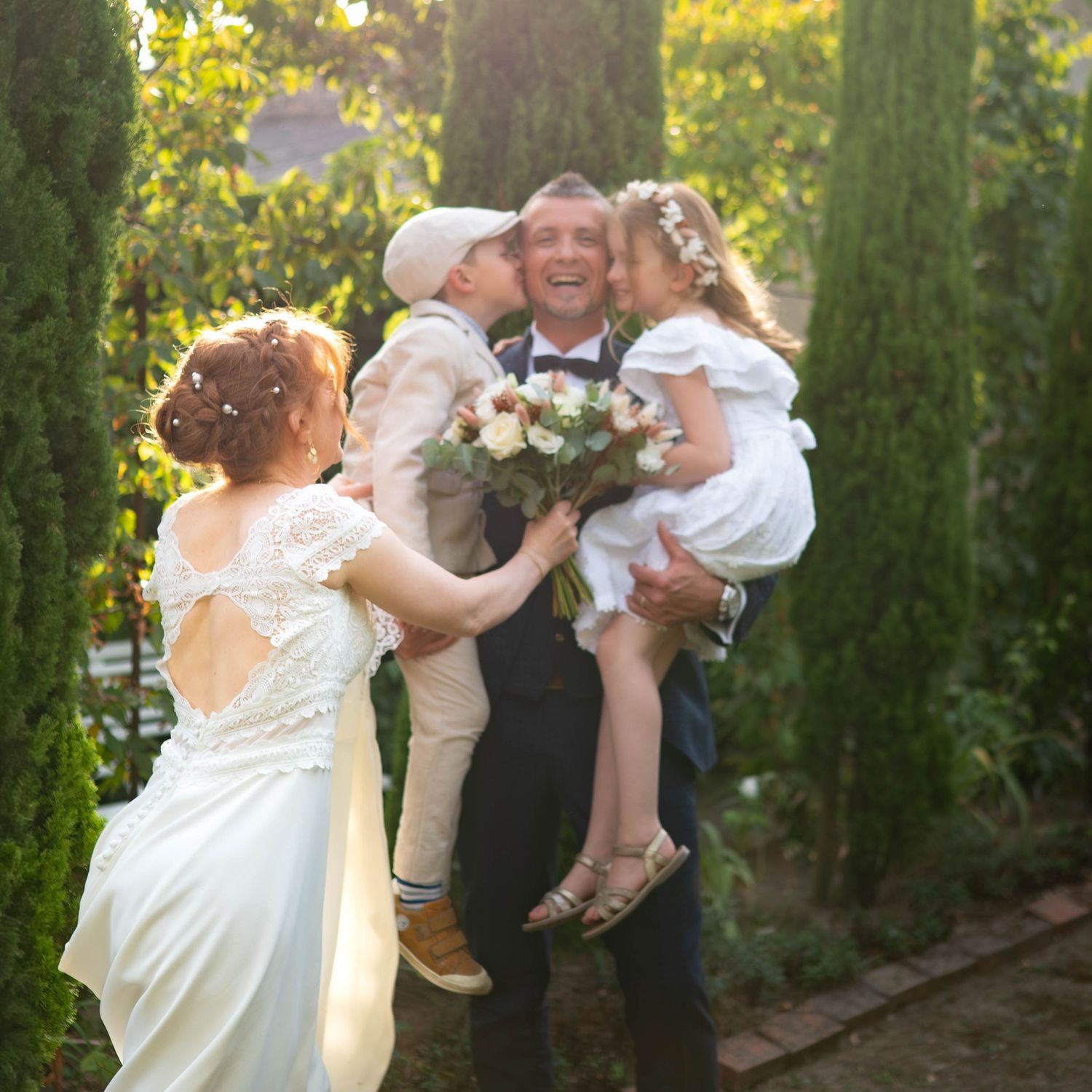 Les mariés dans un jardin, tenant dans leurs bras deux jeunes enfants lors d'une cérémonie de mariage en plein air.