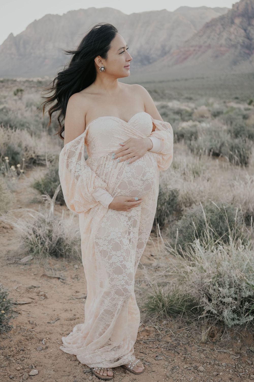 Pregnant woman in elegant lace maternity gown poses outdoors in desert landscape with red rock mountains backdrop.