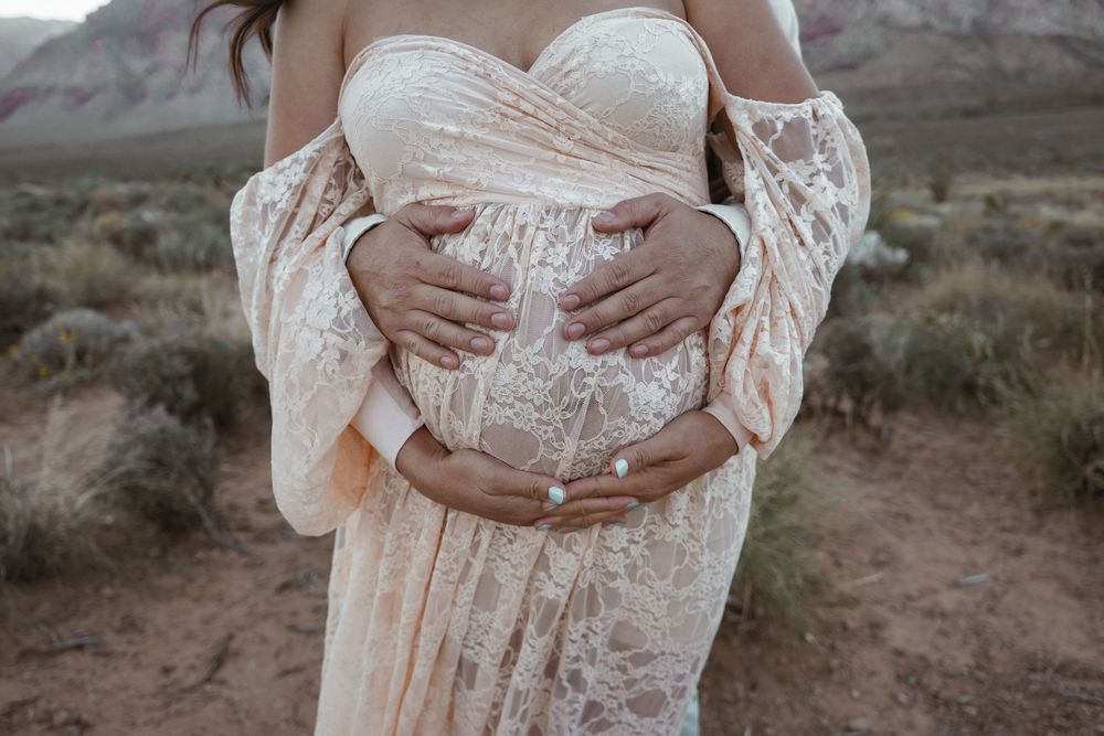 Pregnant woman in elegant lace off-shoulder gown cradling baby bump outdoors in desert landscape.