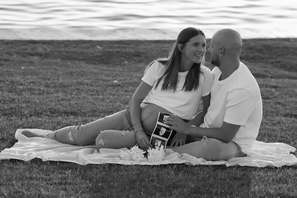 Couple sitting on blanket by water holding ultrasound photo in black and white maternity announcement.