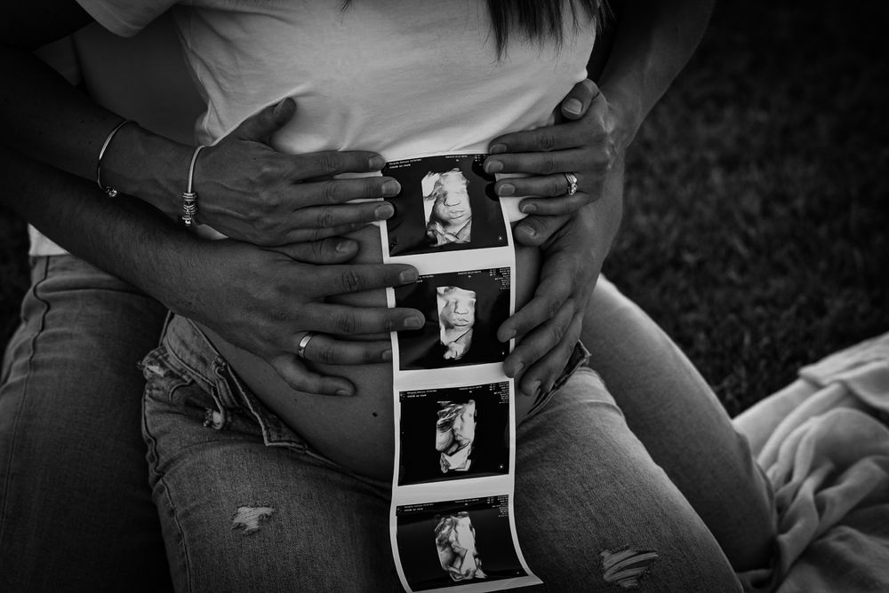 Black and white photo of pregnant couple holding ultrasound strip over baby bump outdoors.