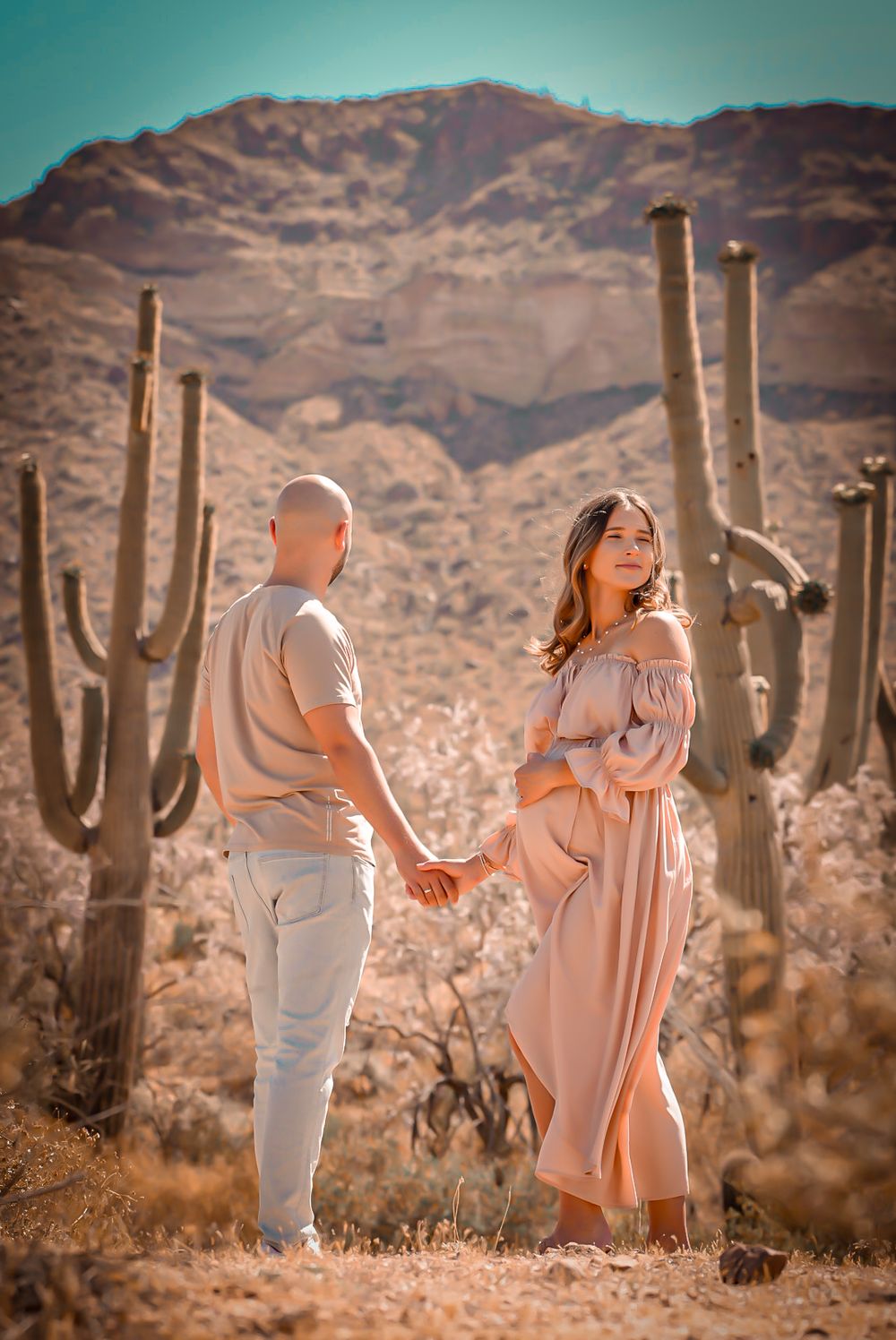 Pregnant couple holding hands in desert with saguaro cacti and mountains in maternity photo shoot.