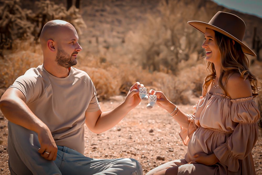 Couple sitting in desert landscape, smiling and clinking small bottles together in a romantic outdoor setting.