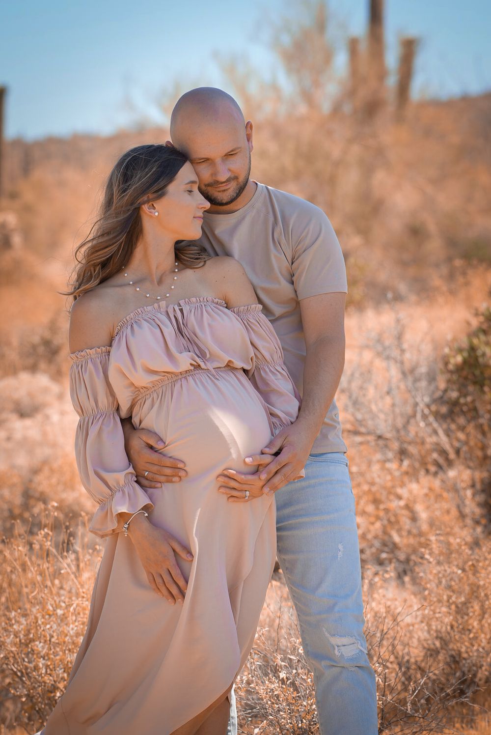 Couple in desert maternity photoshoot, woman in blush off-shoulder gown, man embracing her baby bump among golden brush.