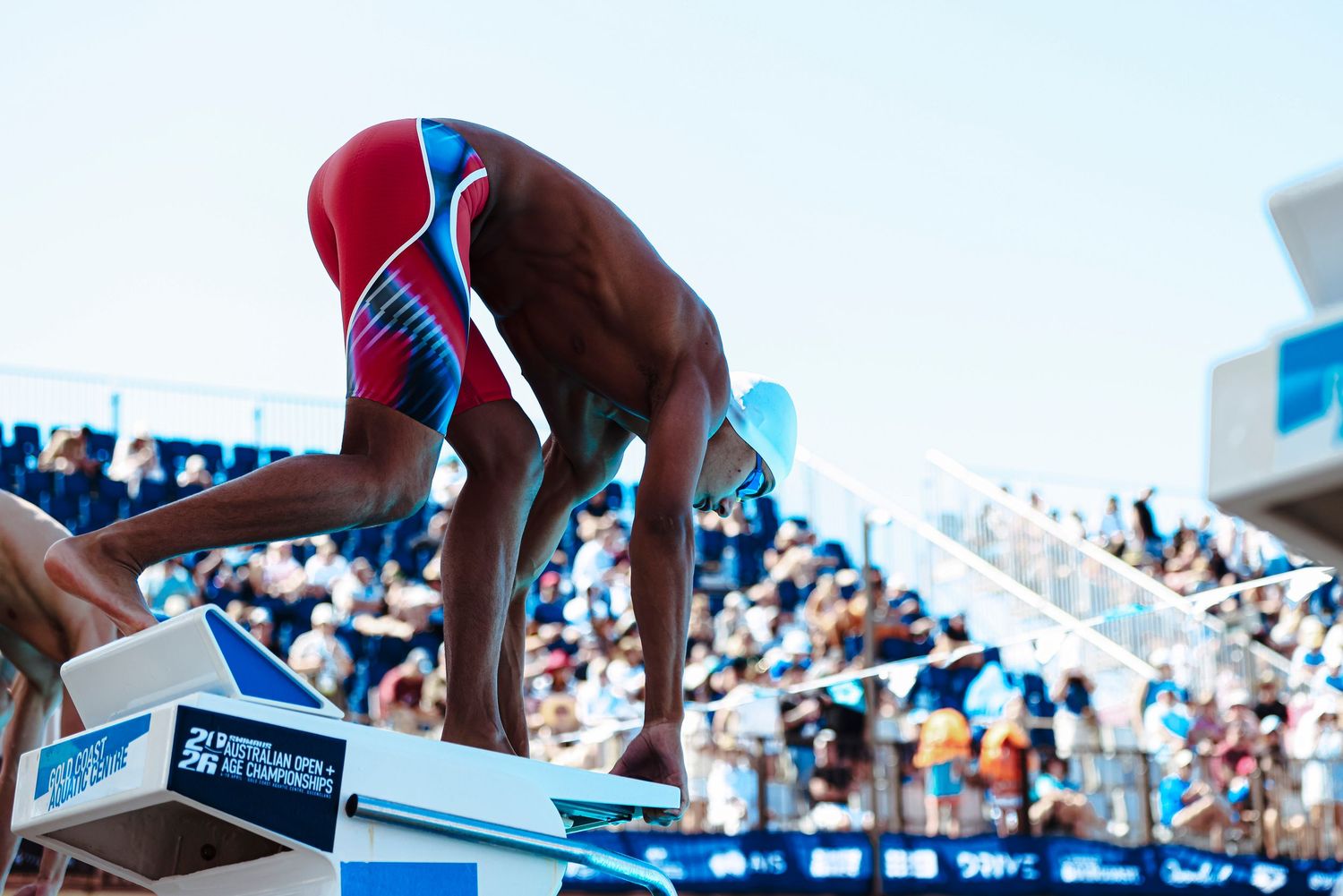 Competitive swimmer in red and blue swimsuit crouches on starting block at an outdoor swim meet with crowded bleachers.