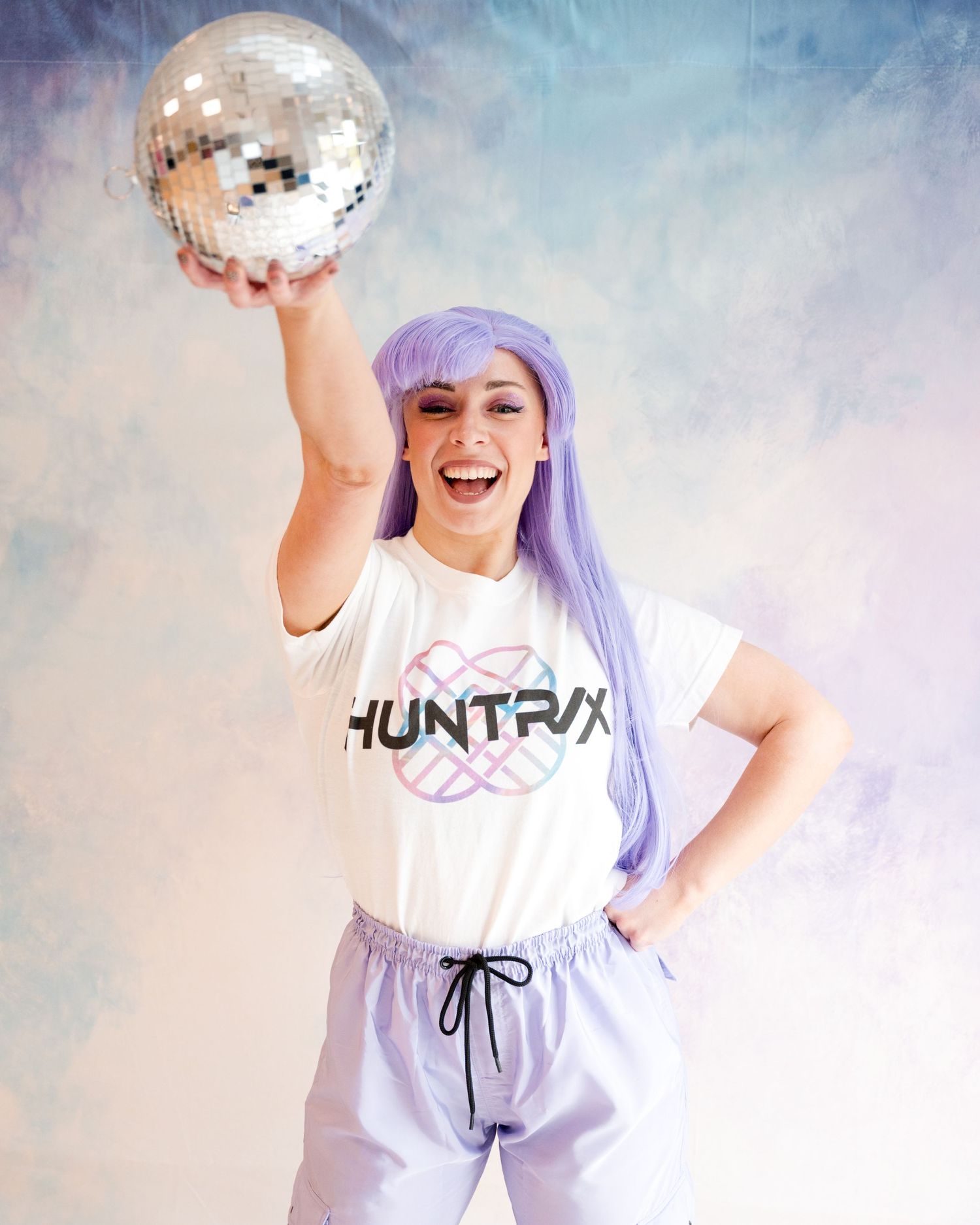 Woman in purple wig and Huntrix shirt holds disco ball overhead while smiling.