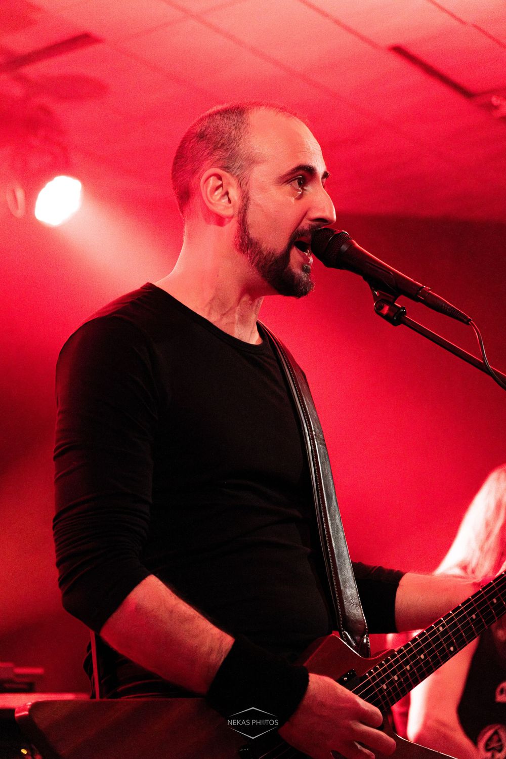 Male vocalist singing into microphone while playing guitar on stage under red lighting at live concert.