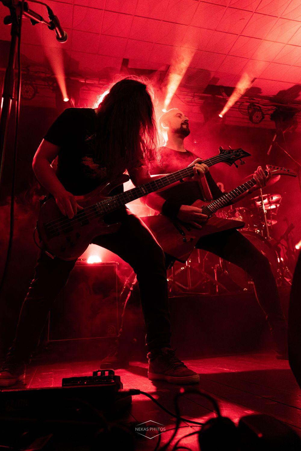 Two guitarists perform on stage bathed in dramatic red lighting during an intense heavy metal concert.