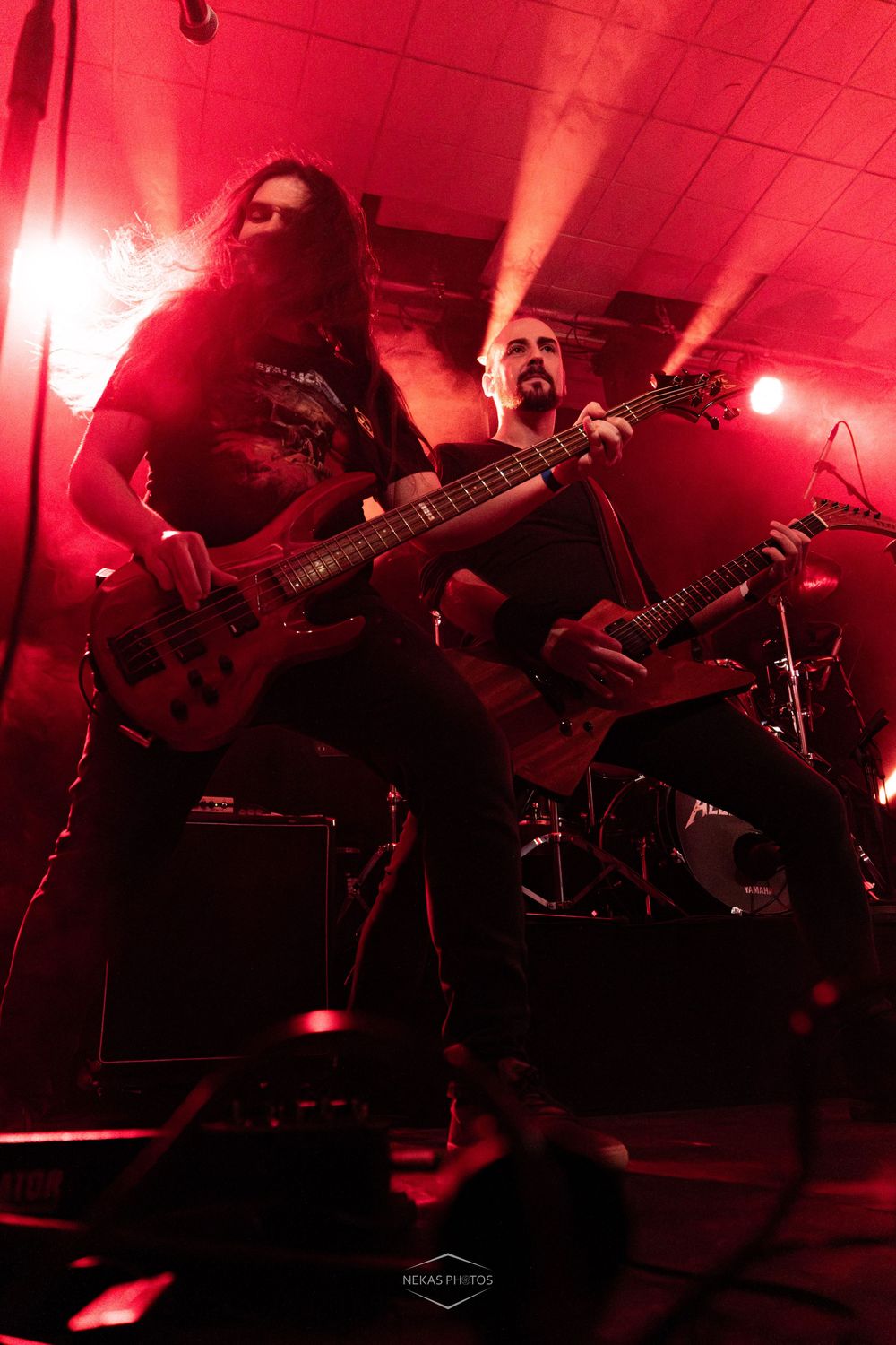 Two metal guitarists perform on a red-lit stage, playing electric guitars with long hair and dark clothing.