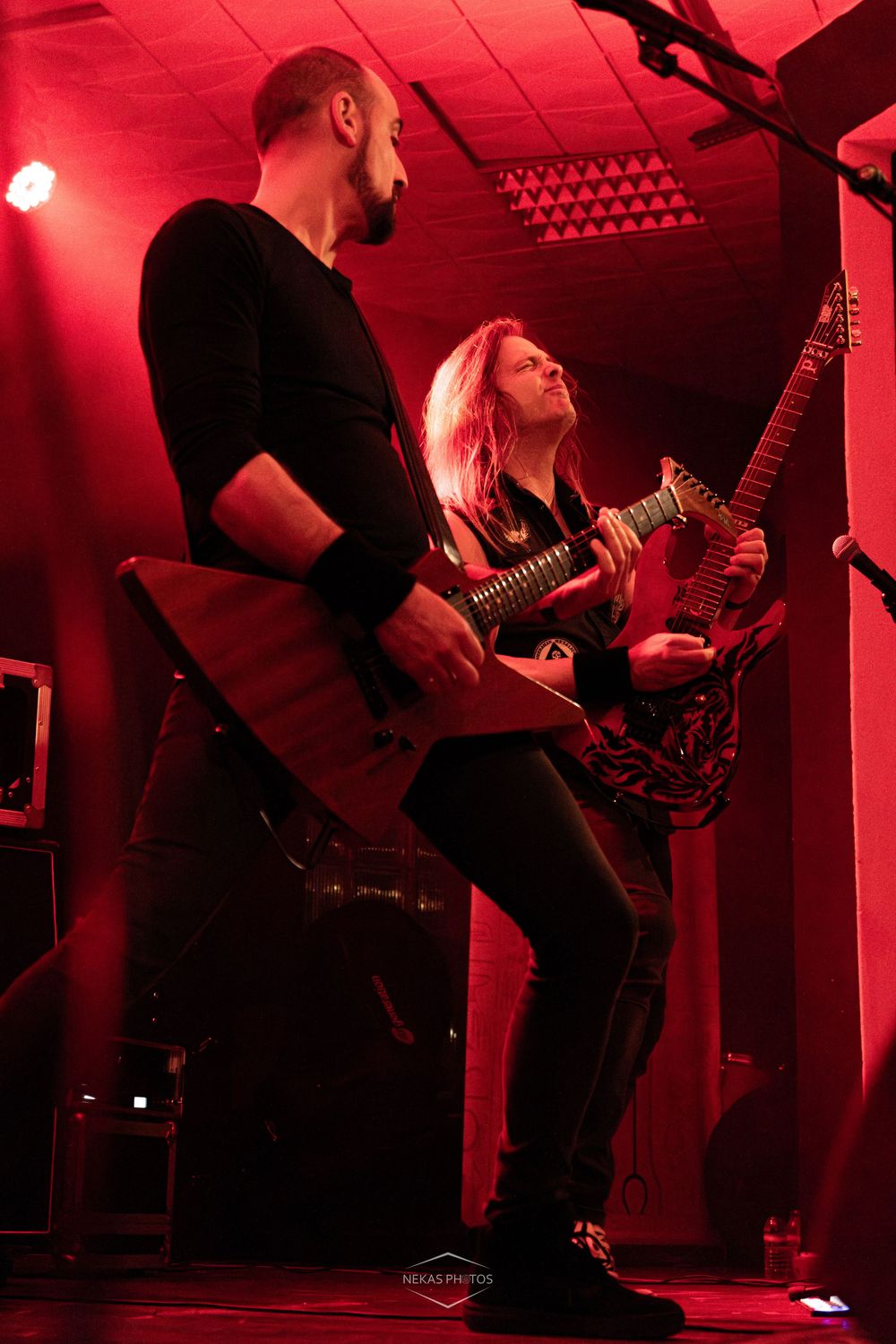 Two guitarists perform on stage under red lights, playing electric V-shaped guitars at a heavy metal concert.