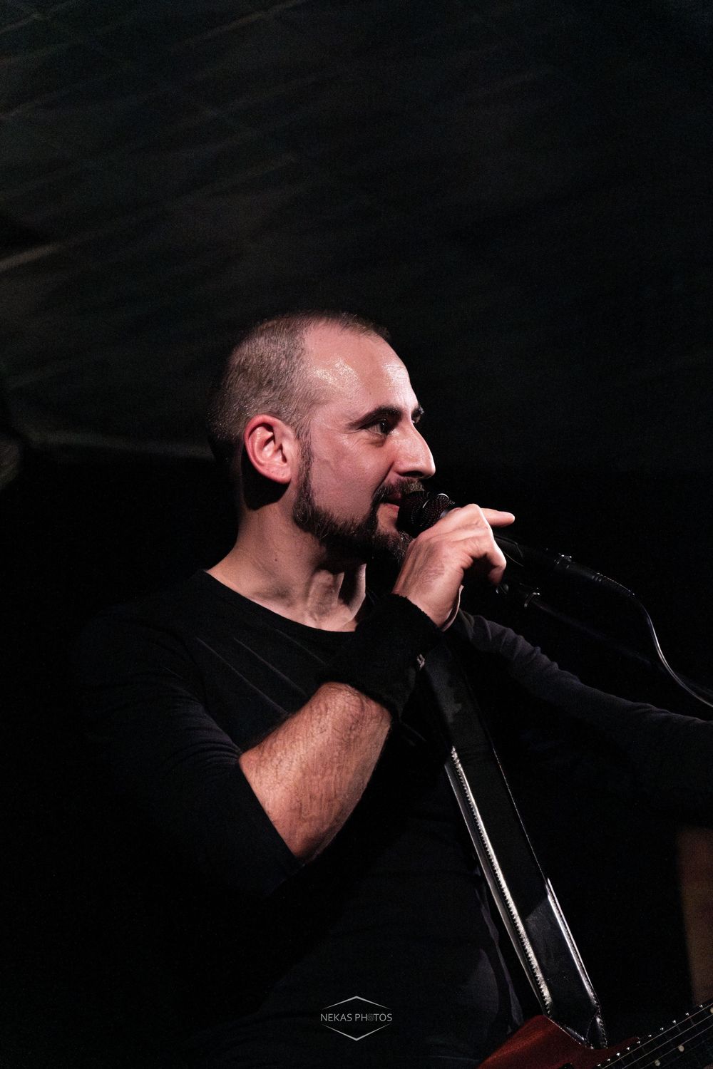 A bearded man in a black shirt performs on stage, singing into a microphone under dramatic lighting.