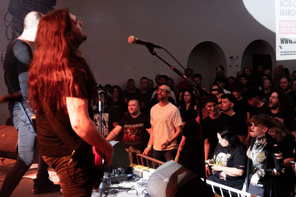 Metal band performing live on stage with long-haired guitarist facing large crowd at indoor music venue.