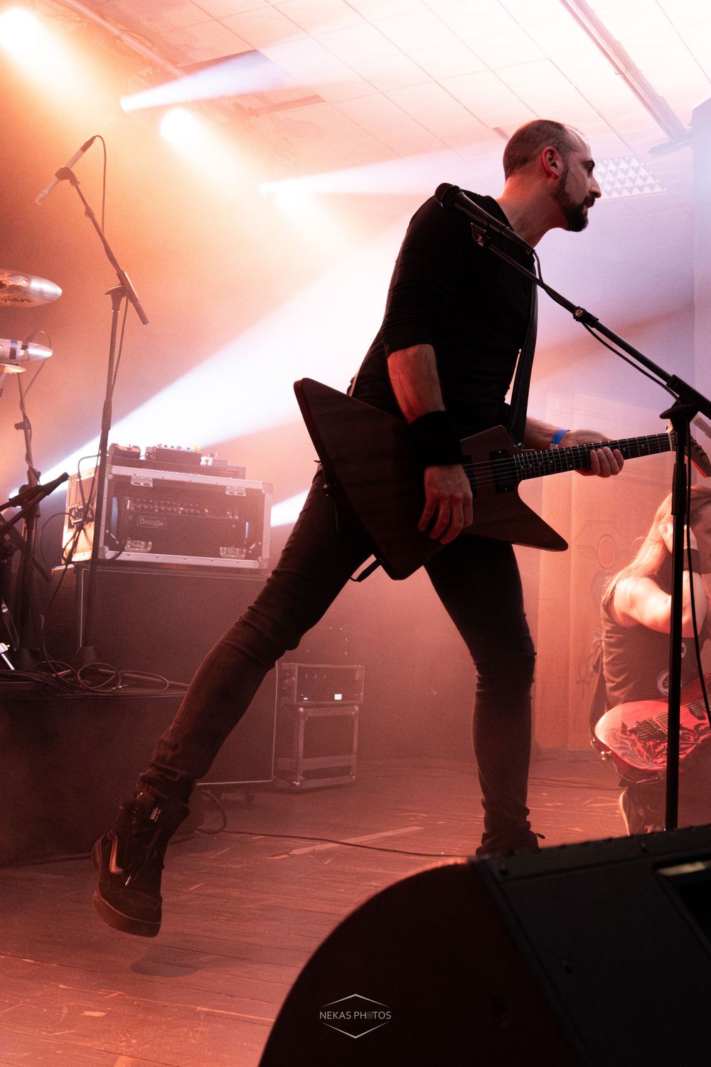 A guitarist jumps on stage during a rock concert, playing a black electric guitar under dramatic orange stage lighting.