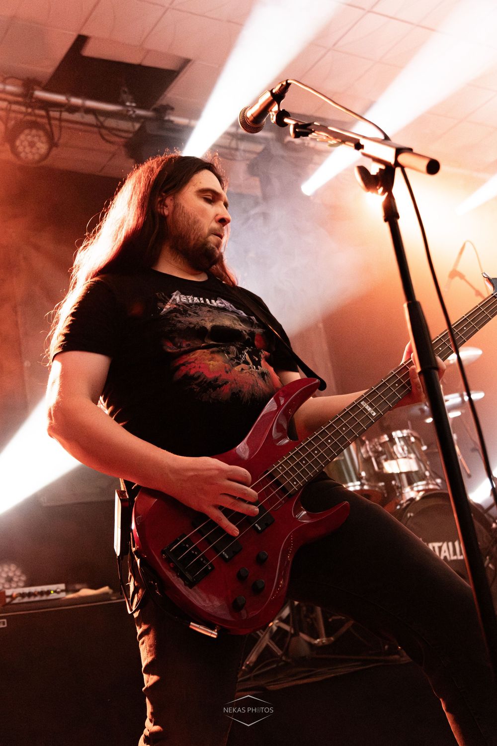 Long-haired male bassist playing red electric bass guitar on stage under dramatic red lighting at a metal concert.