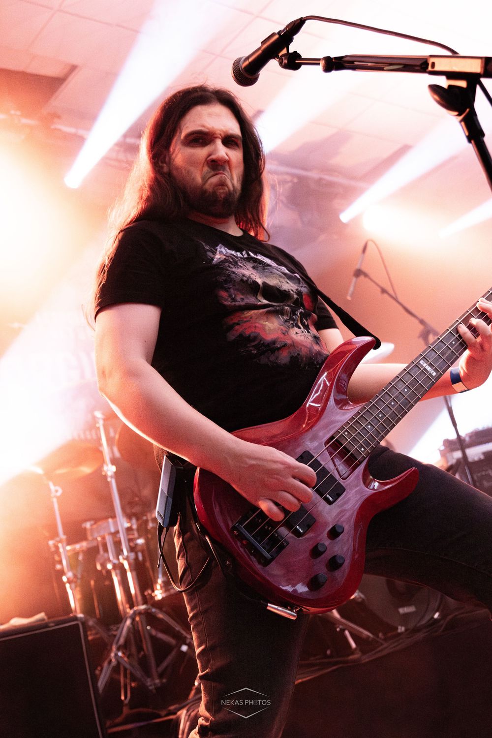 Long-haired male guitarist playing a red electric guitar on stage under bright lights at a live metal concert.
