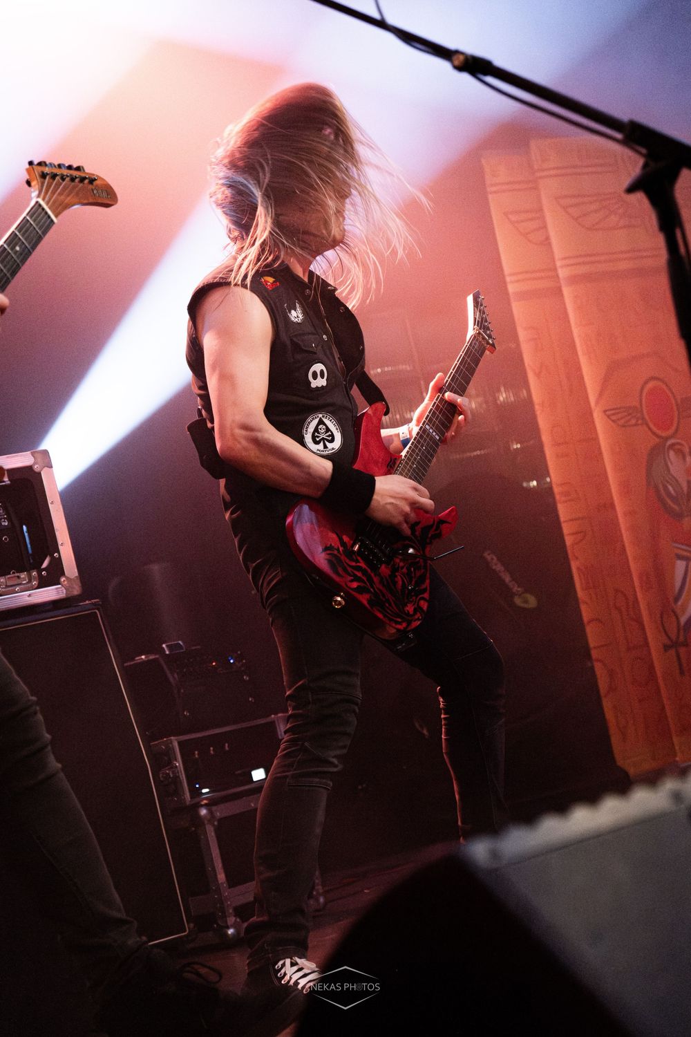 A guitarist headbanging on stage, playing a red electric guitar, wearing a sleeveless vest under dramatic pink and orange stage lighting.