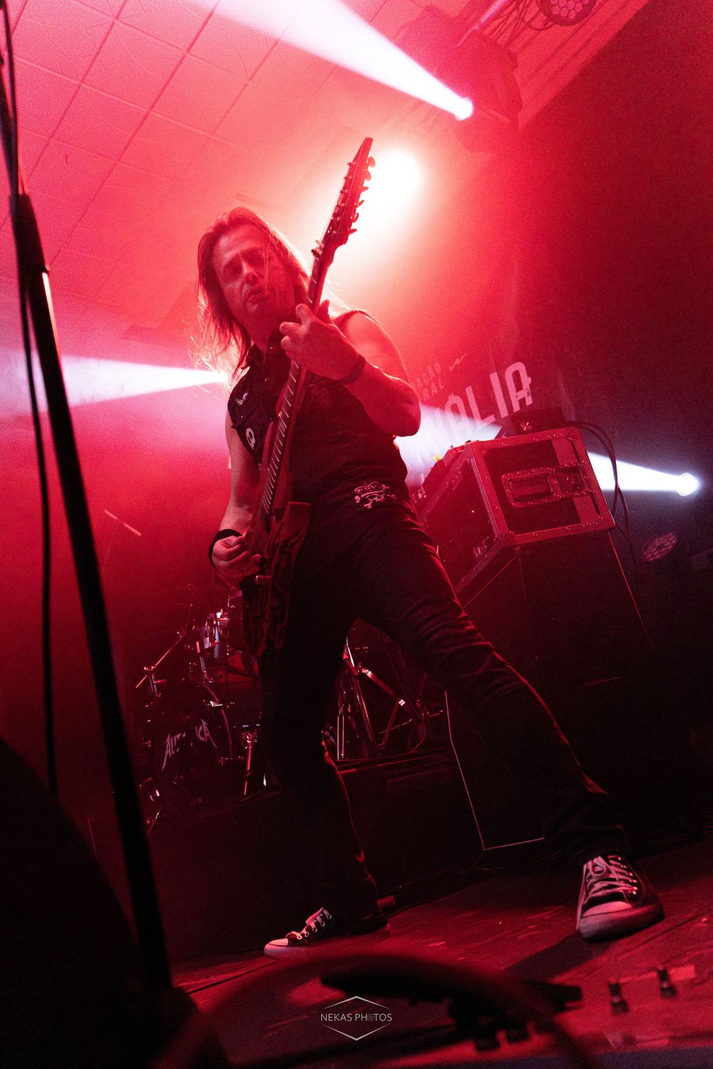 Metal guitarist performs on stage bathed in red light, playing electric guitar with long hair and black outfit.