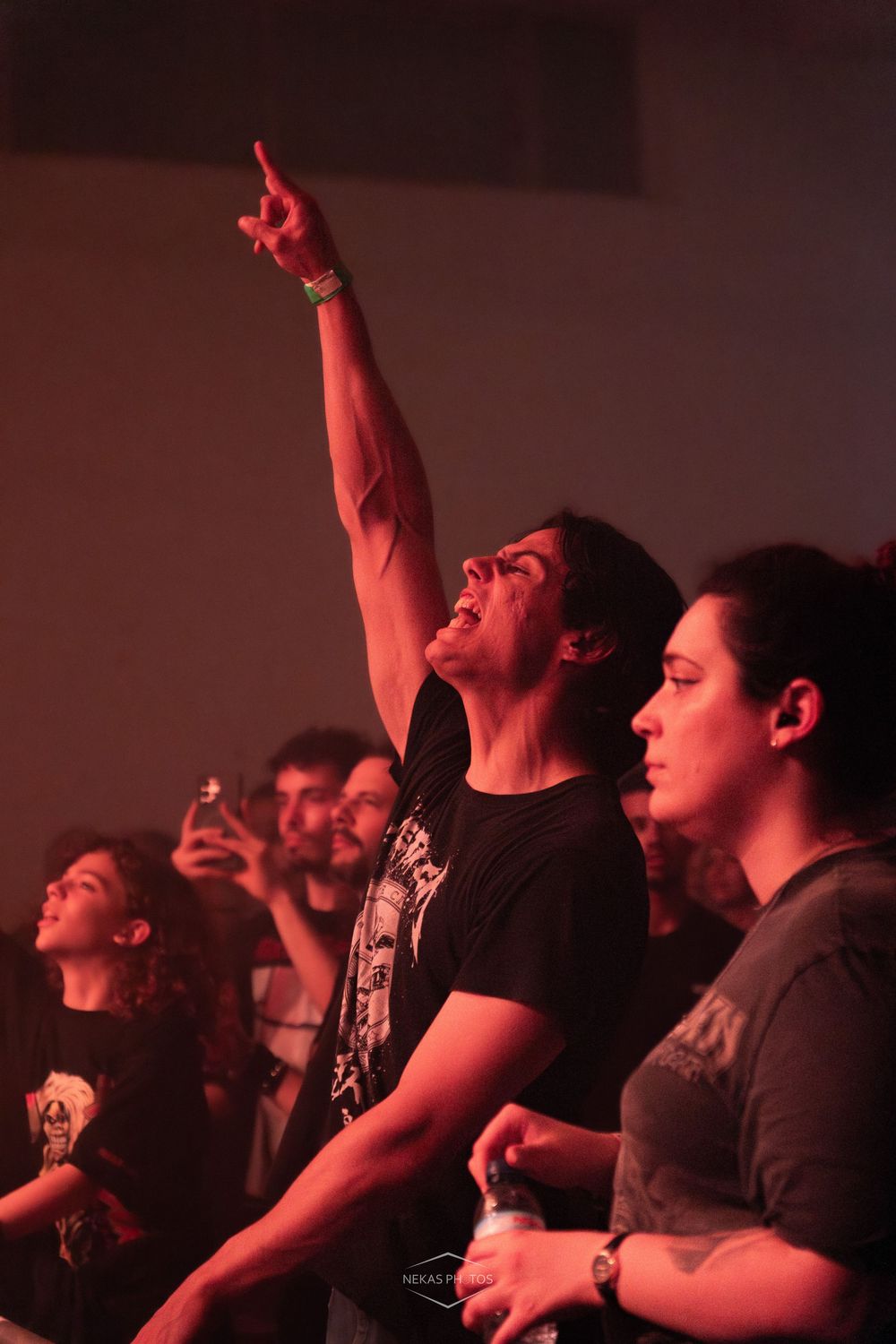 Excited concert-goer points upward with raised arm in red-lit venue crowd during live music event.