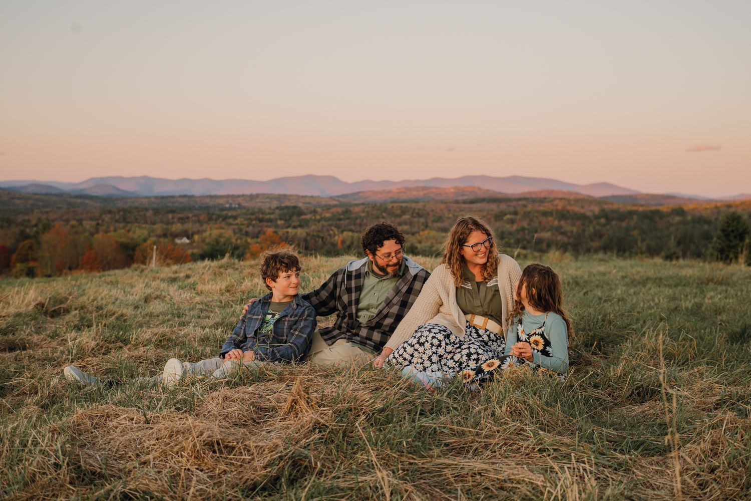 Family of four sits together on a hay bale in a golden field with mountain views at sunset.