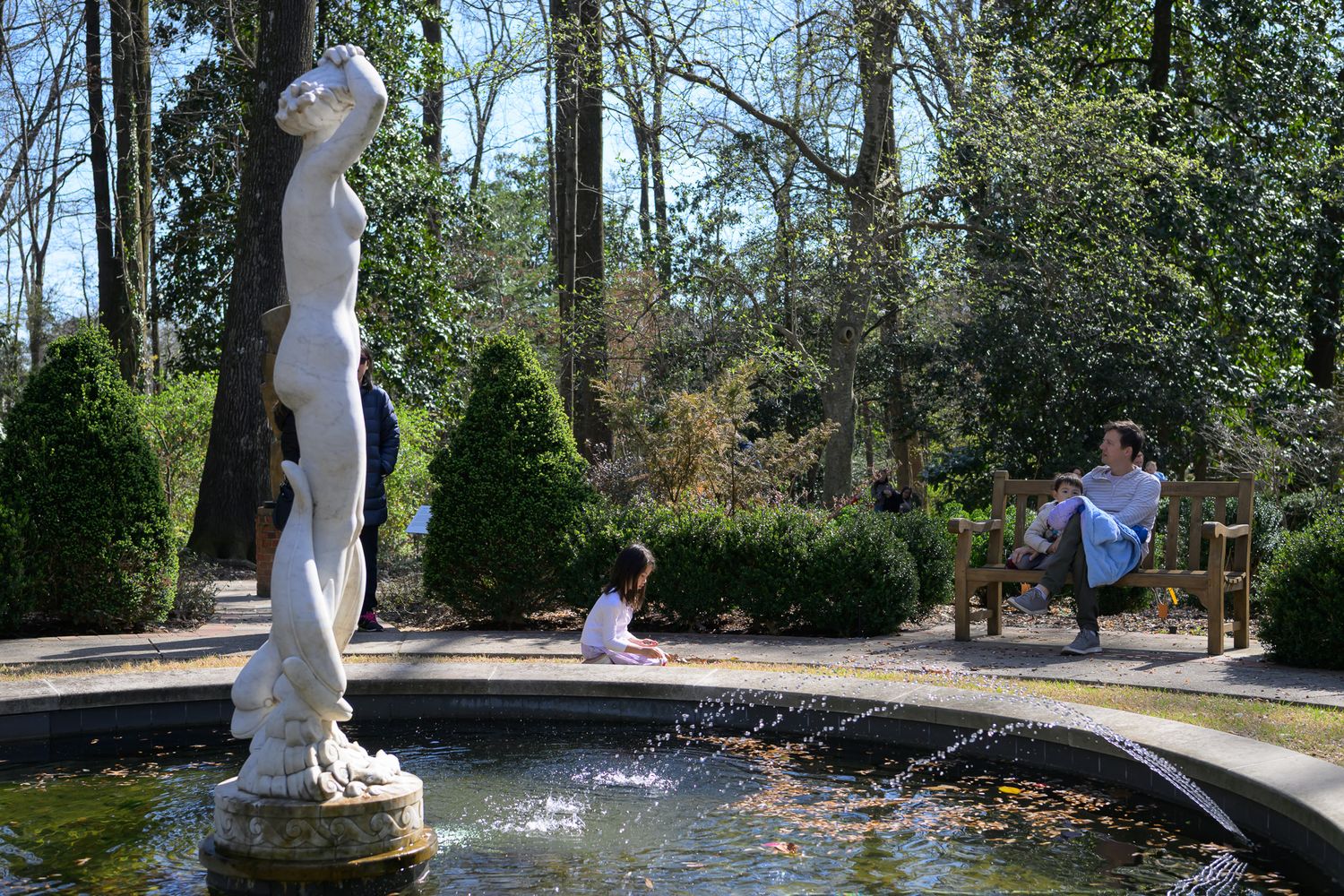 Classical stone fountain statue in a sunlit garden with children playing nearby and lush green hedges in the background.
