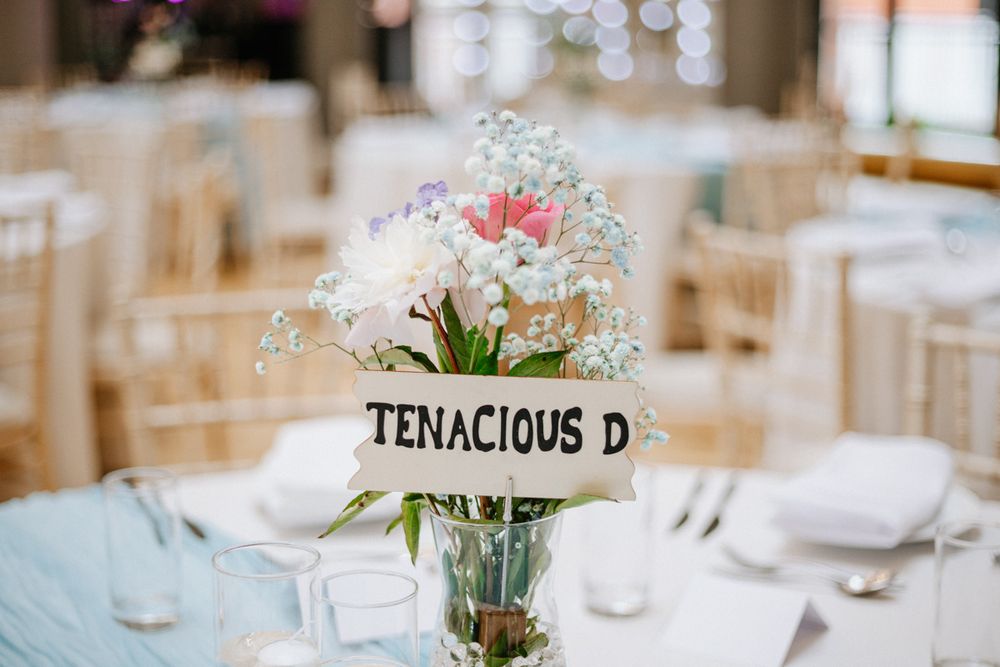 Wedding reception table centerpiece with pink flowers and baby's breath in a vase, featuring a Tenacious D table name sign.