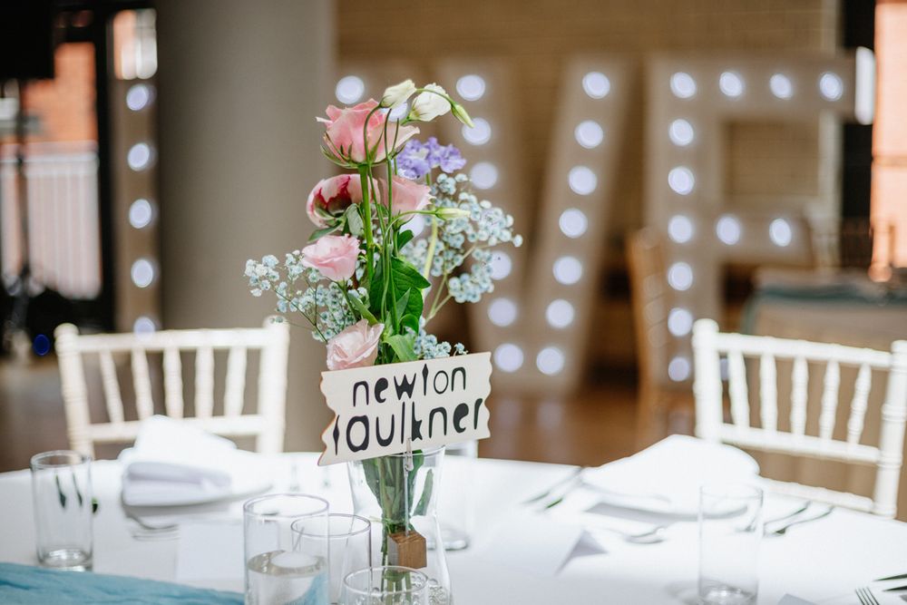 Elegant wedding table centerpiece with pink flowers in a glass vase, Newton Faulkner name card, white chairs, and lit LOVE letters.