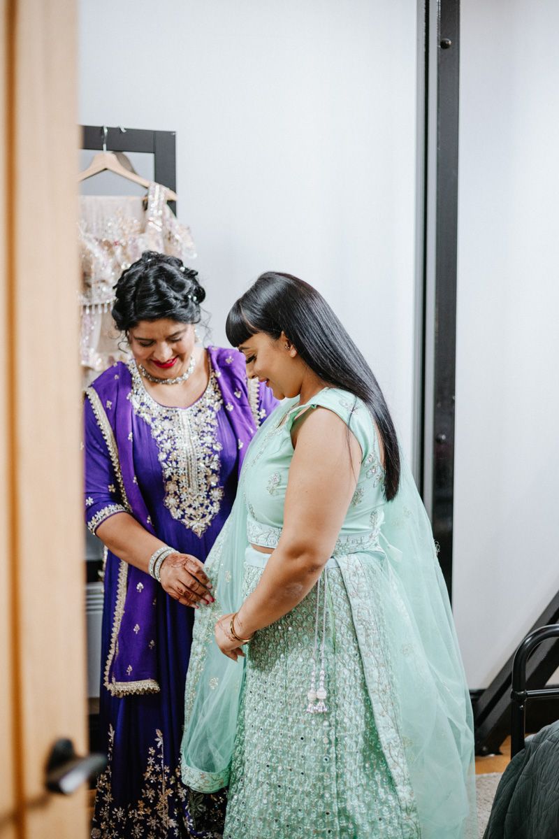 Two women in traditional Indian attire, one in purple salwar and one in mint green lehenga, sharing a joyful moment.
