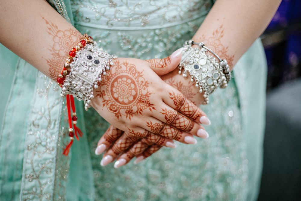 Close-up of bride's hands adorned with intricate henna mehndi design and silver bangles on mint green lehenga.