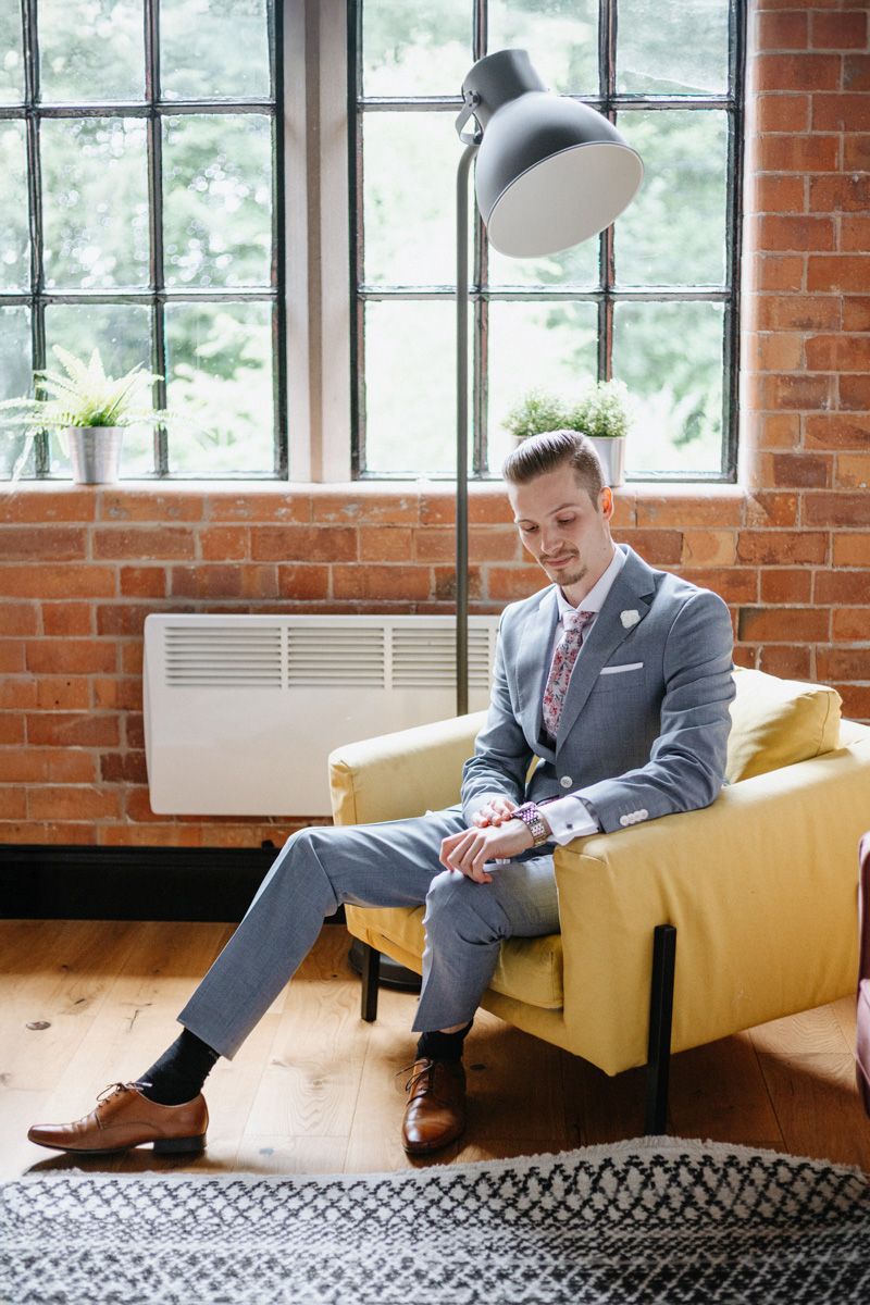 Stylish groom in grey suit sitting on yellow armchair in modern loft with brick wall and large windows.