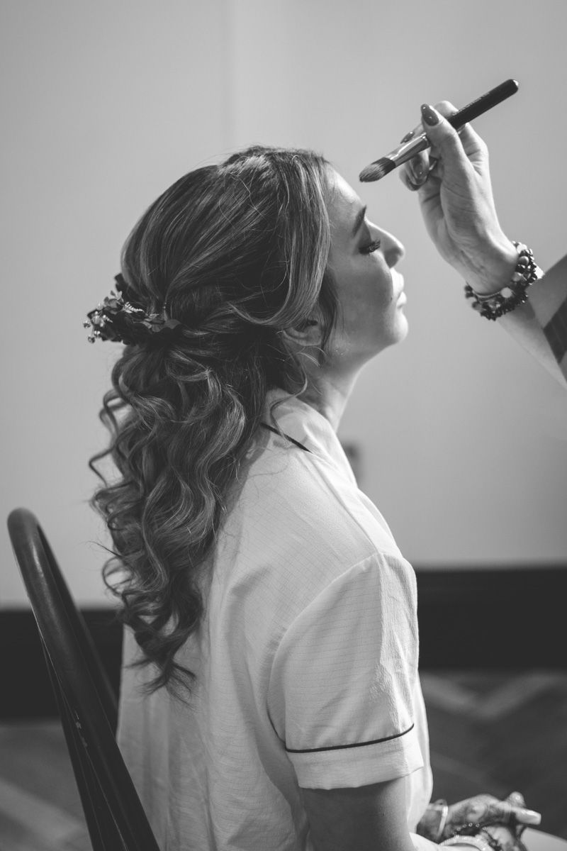 Black and white photo of a bride getting makeup applied, featuring elegant curly hair adorned with small flowers.