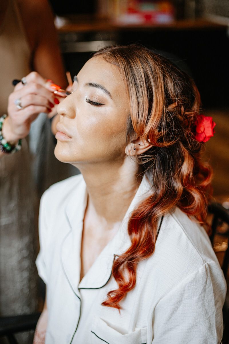 Bride getting makeup applied with red flower hair accessory and curled auburn hair in white robe.
