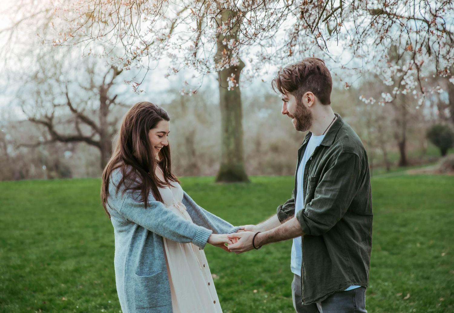 A young couple holds hands tenderly in a lush green park surrounded by blooming trees in spring.