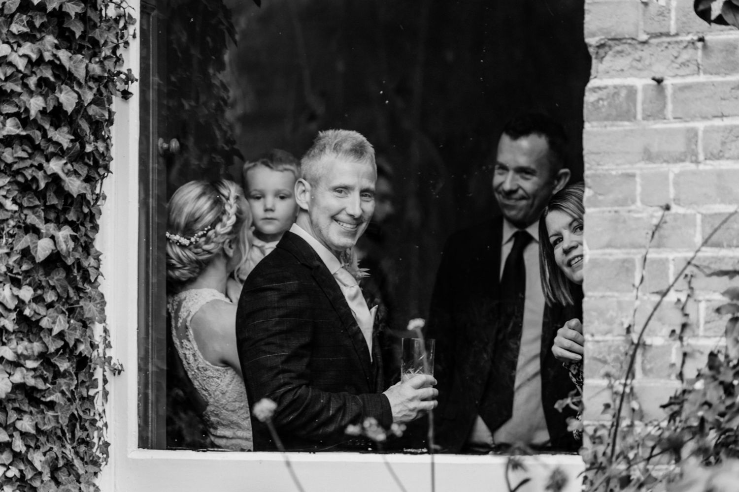 Black and white photo of smiling wedding guests peering through a window framed by ivy-covered brick walls.