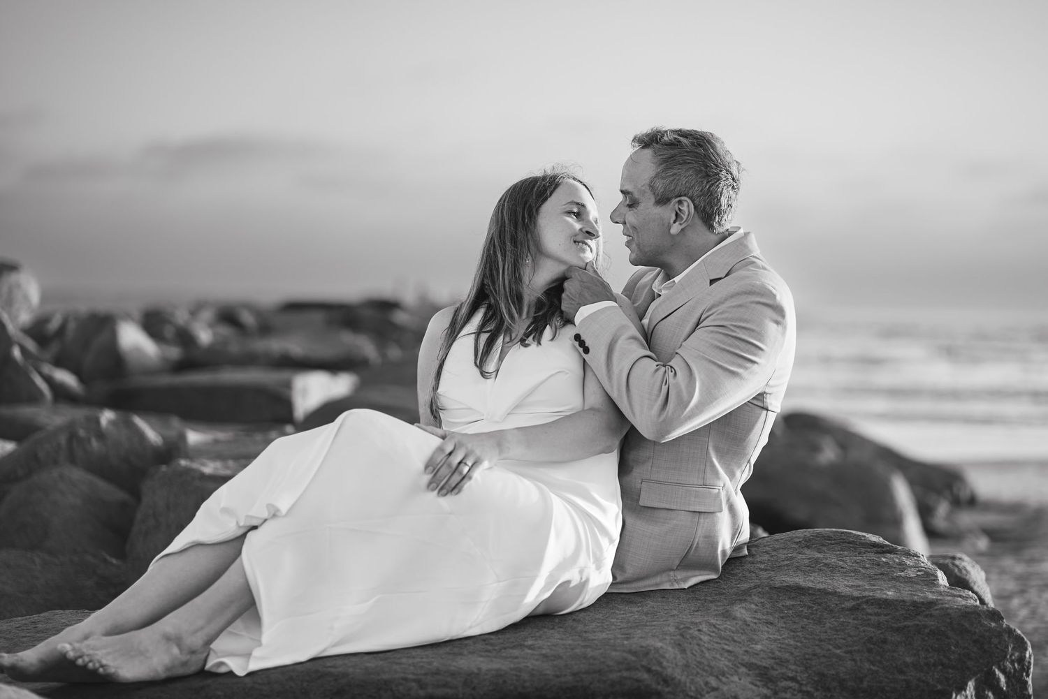 Black and white photo of a newly married couple sitting on rocks by the ocean, gazing lovingly at each other. Oceanside, CA