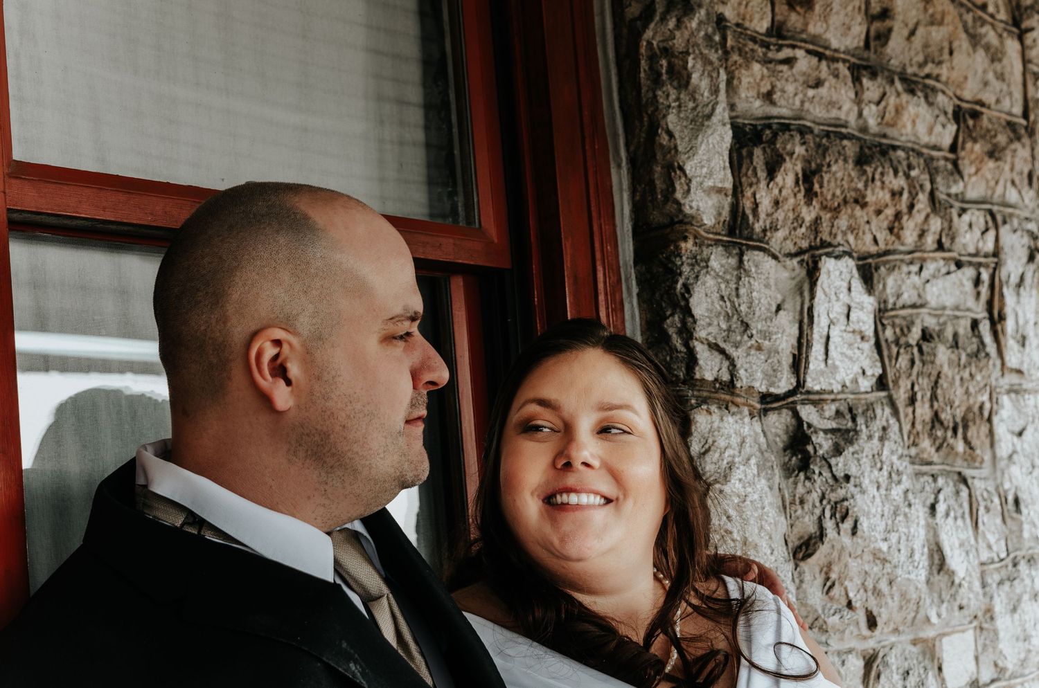 A smiling couple in wedding attire poses near a rustic stone wall and red window frame.