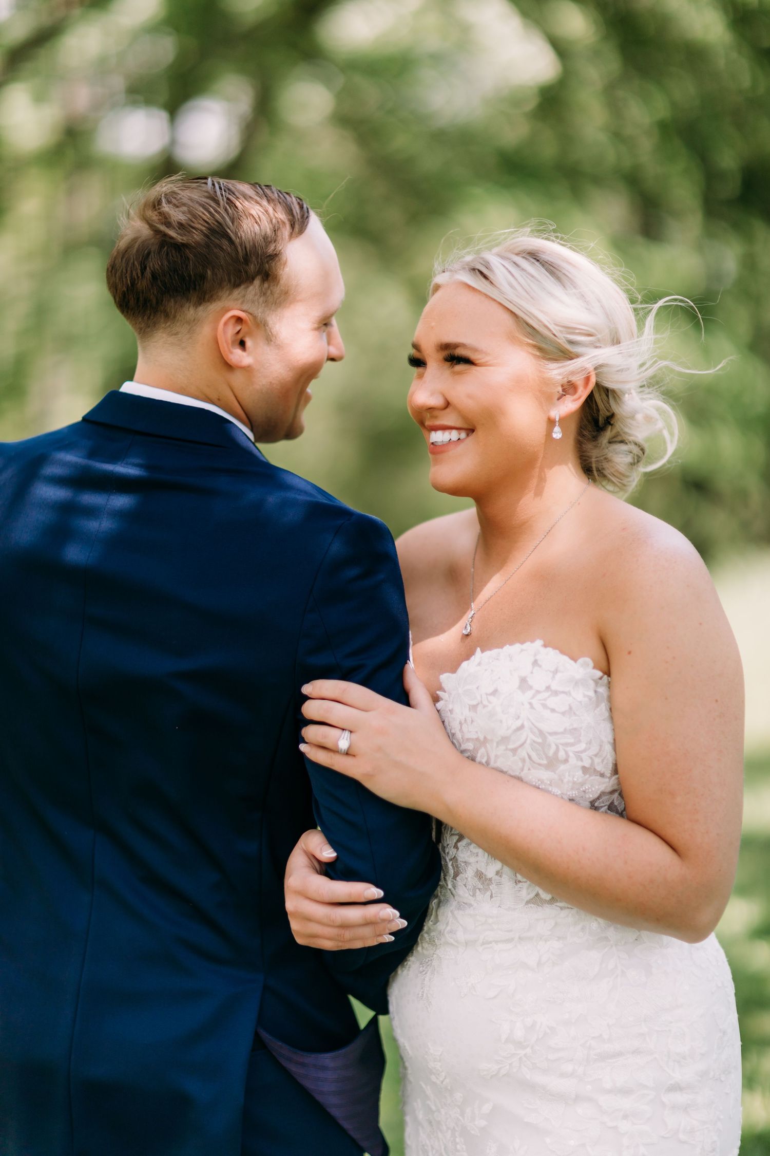 Bride in strapless lace gown smiles at groom in navy suit during outdoor wedding portrait.