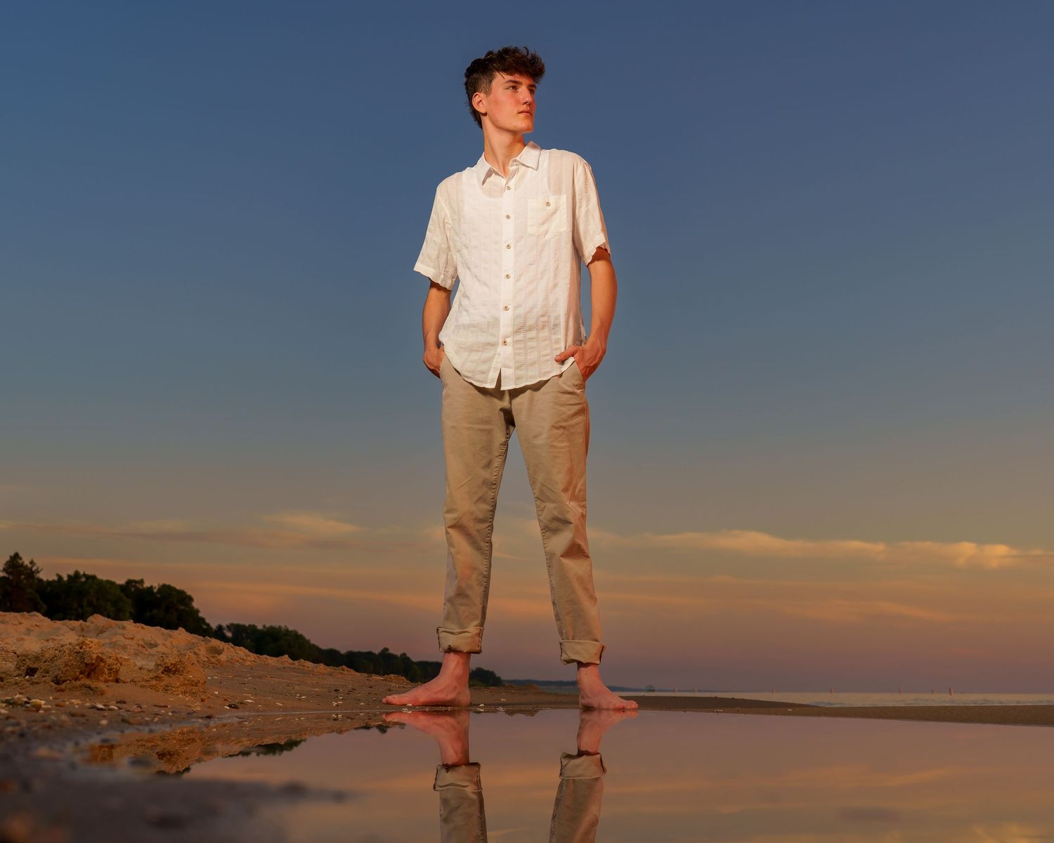 A barefoot teenage boy in white shirt and khaki pants stands on a reflective beach at sunset.