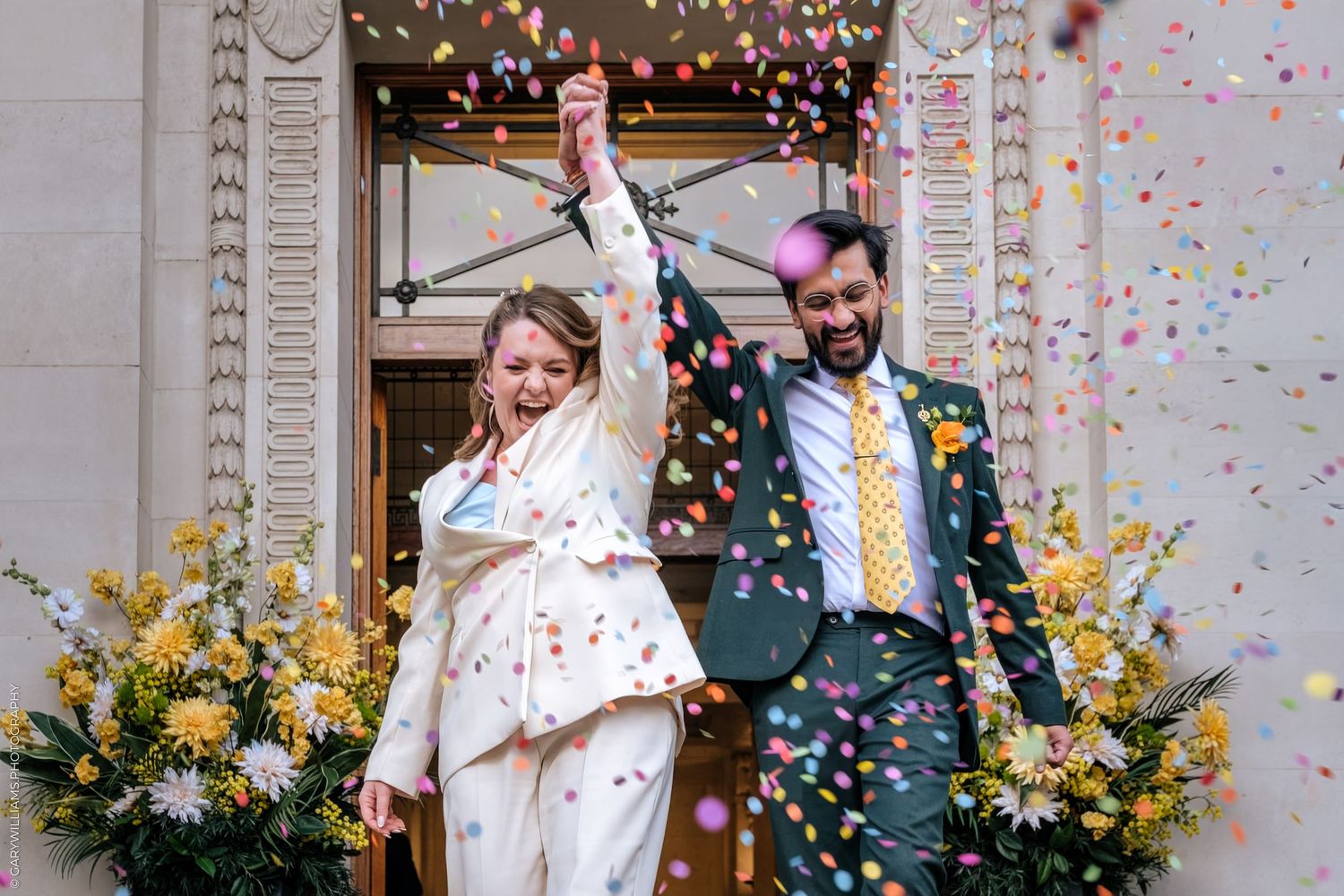 Joyful newlyweds celebrate outside with colorful confetti, bride in white suit and groom in dark suit with yellow tie.