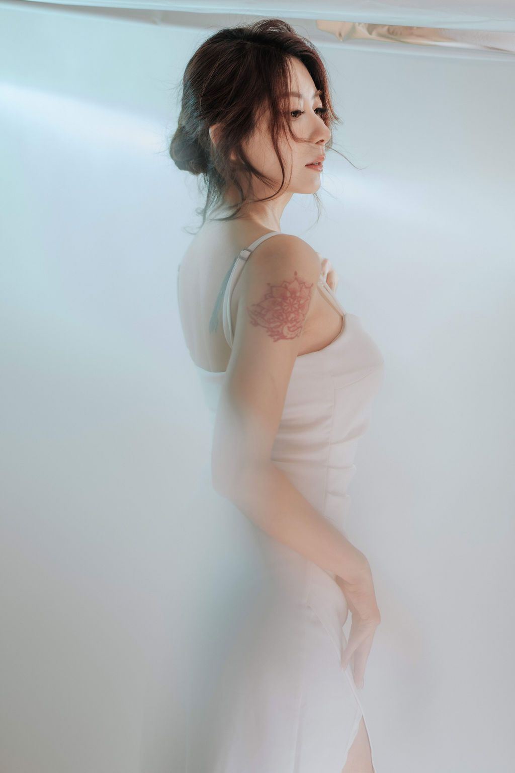 Woman in white dress with floral shoulder tattoo poses elegantly against a bright white background.