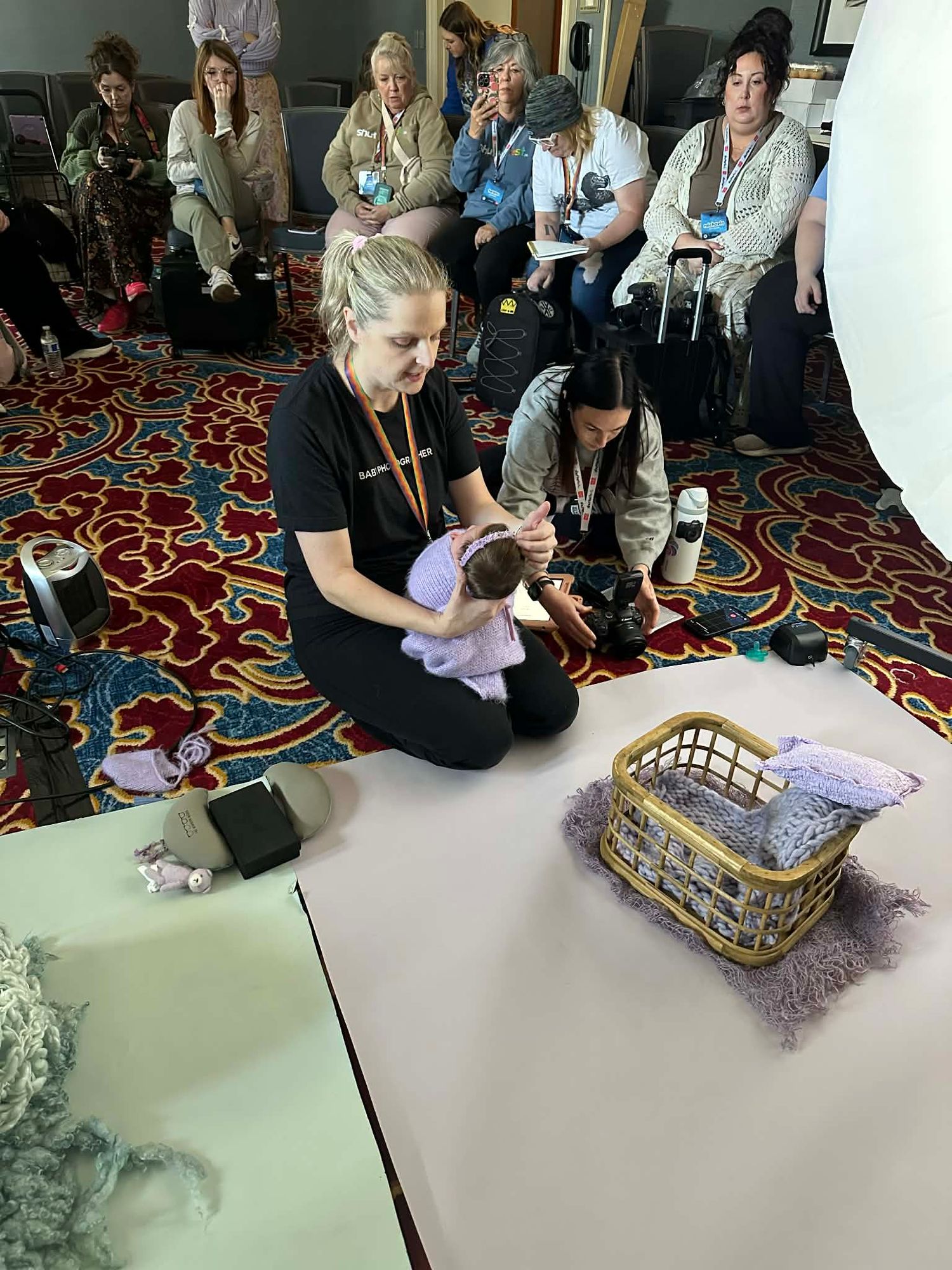 Women sit on patterned carpet during a newborn photography workshop, posing a baby doll in a wicker basket prop.
