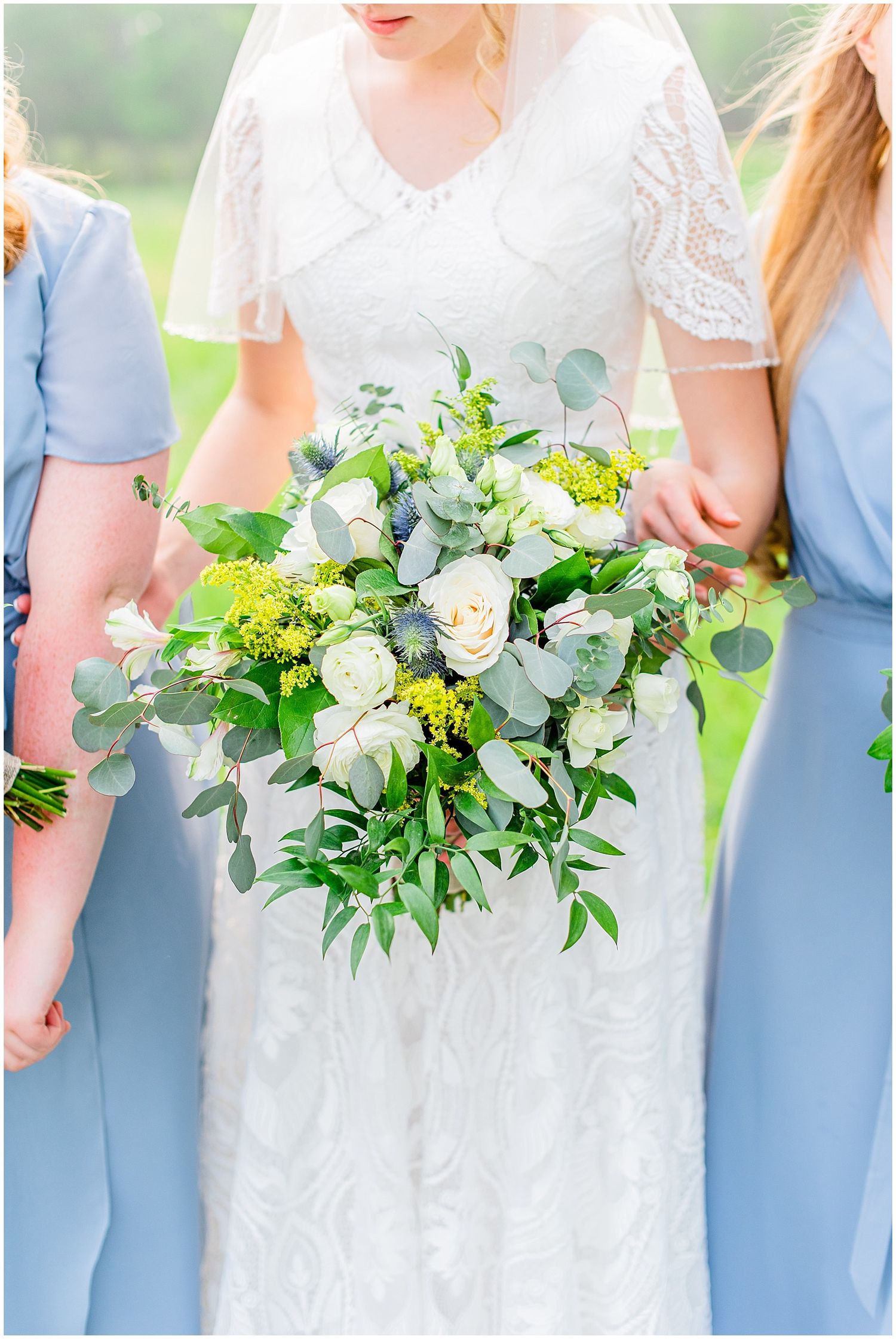 Bride in lace gown holds bouquet of white roses, eucalyptus, and greenery flanked by bridesmaids in blue dresses.