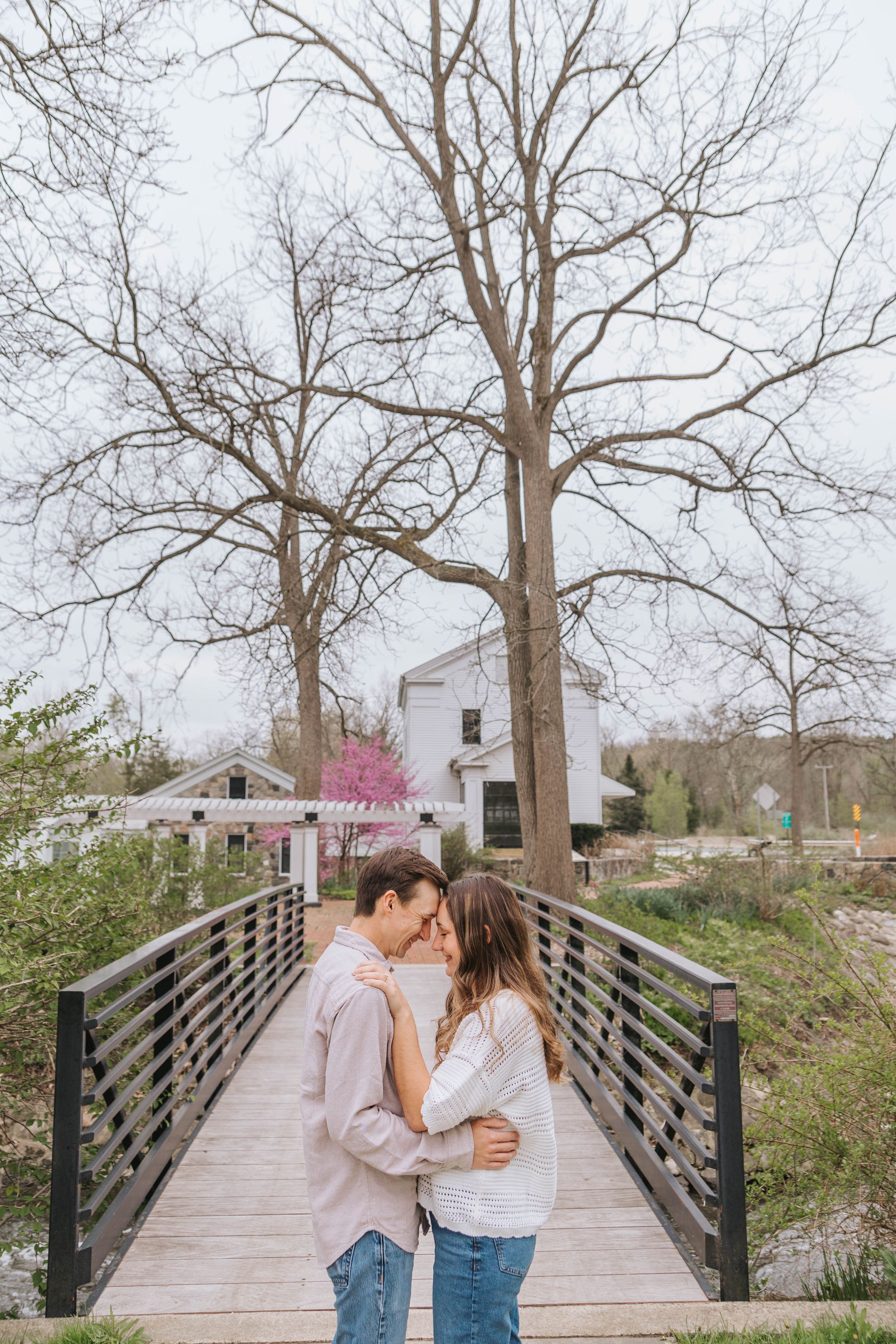 Couple sharing a romantic moment on a wooden bridge surrounded by bare trees and a white building in early spring.
